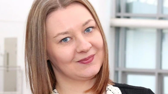 A woman with shoulder-length blonde hair and blue eyes is wearing a black dress with a white lace collar. She is smiling subtly in a bright indoor setting with large windows behind her. Arlene Sweeney Learner Engagement Officer